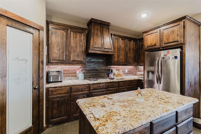 kitchen with a kitchen island, custom range hood, appliances with stainless steel finishes, light stone countertops, and dark brown cabinets