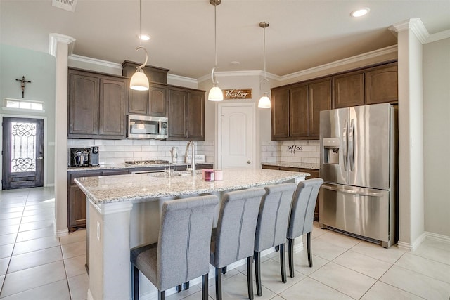 kitchen with dark brown cabinetry, light stone counters, light tile patterned floors, appliances with stainless steel finishes, and a kitchen island with sink