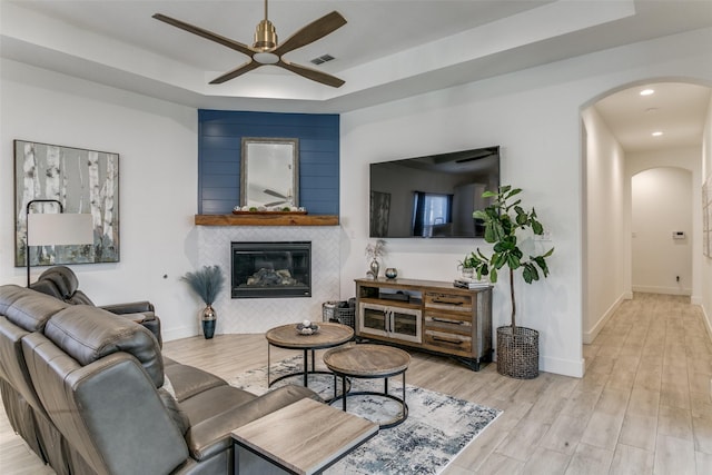 living room featuring ceiling fan, a raised ceiling, a tile fireplace, and light wood-type flooring