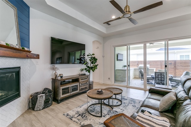 living room with a tiled fireplace, ceiling fan, a raised ceiling, and light hardwood / wood-style floors