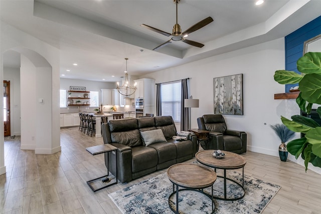 living room with a raised ceiling, ceiling fan with notable chandelier, and light hardwood / wood-style flooring