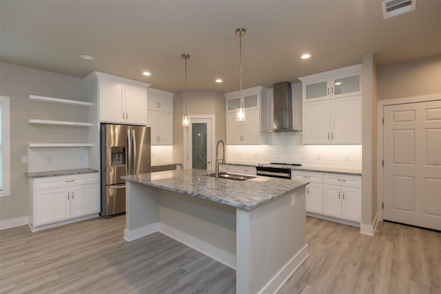 kitchen featuring wall chimney range hood, sink, white cabinetry, stainless steel appliances, and an island with sink