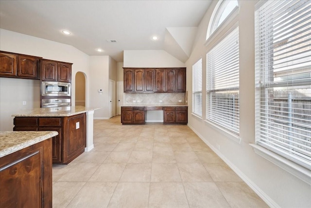 kitchen featuring dark brown cabinetry, lofted ceiling, light stone counters, appliances with stainless steel finishes, and backsplash