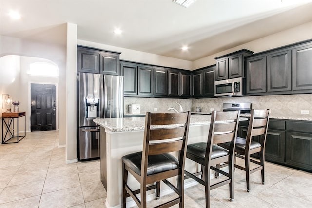 kitchen with arched walkways, light stone counters, appliances with stainless steel finishes, a kitchen breakfast bar, and backsplash