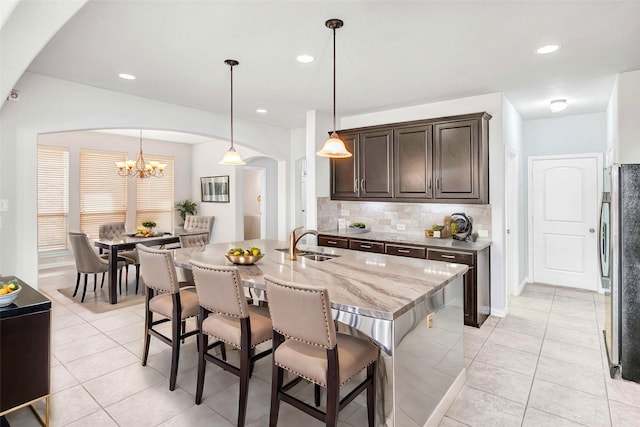 kitchen with light tile patterned flooring, dark brown cabinetry, sink, decorative light fixtures, and a center island with sink