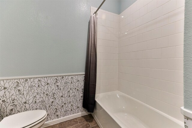 bathroom featuring wallpapered walls, baseboards, a wainscoted wall, toilet, and shower / tub combo