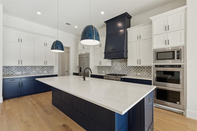 kitchen with a kitchen island with sink, built in appliances, white cabinets, and decorative light fixtures