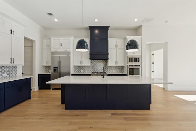 kitchen featuring built in appliances, an island with sink, hanging light fixtures, and light hardwood / wood-style flooring