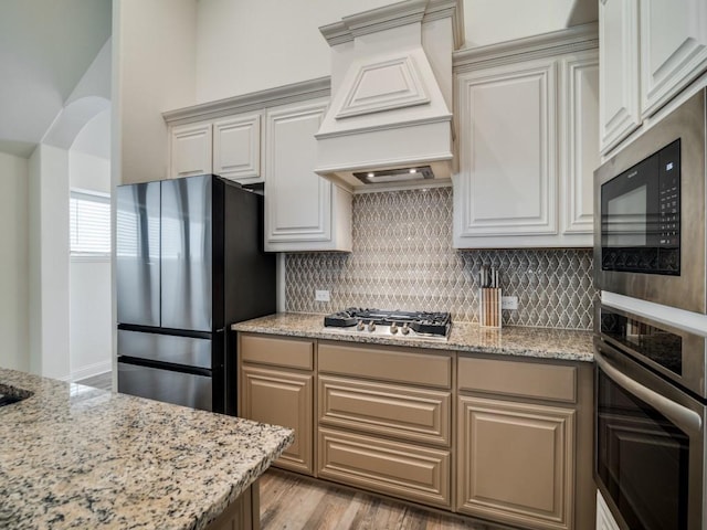 kitchen with stainless steel appliances, light stone countertops, light hardwood / wood-style floors, decorative backsplash, and custom exhaust hood