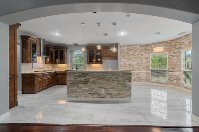 kitchen with pendant lighting, wall chimney range hood, backsplash, stainless steel gas cooktop, and a kitchen island