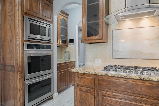 kitchen featuring light stone counters, stainless steel appliances, decorative backsplash, and wall chimney range hood