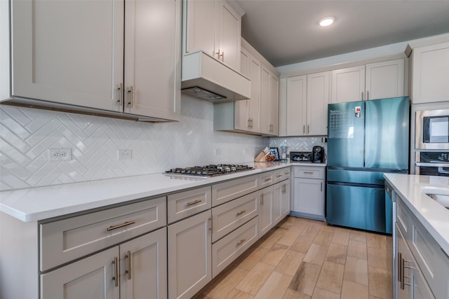 kitchen featuring gray cabinets, stainless steel appliances, decorative backsplash, custom exhaust hood, and light wood-type flooring