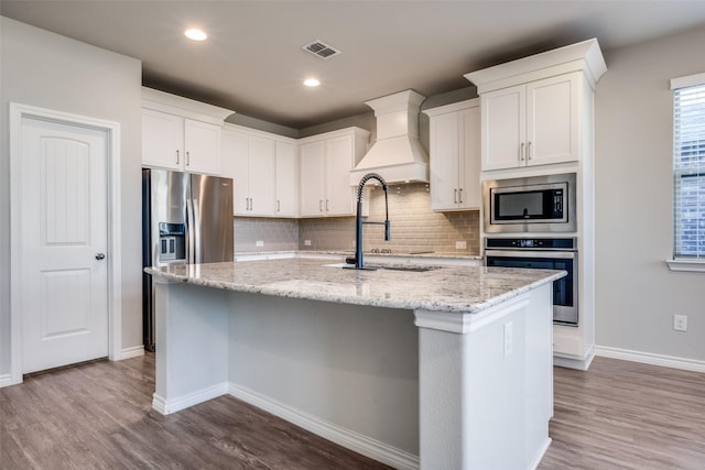 kitchen featuring a kitchen island with sink, stainless steel appliances, light stone countertops, white cabinets, and custom exhaust hood