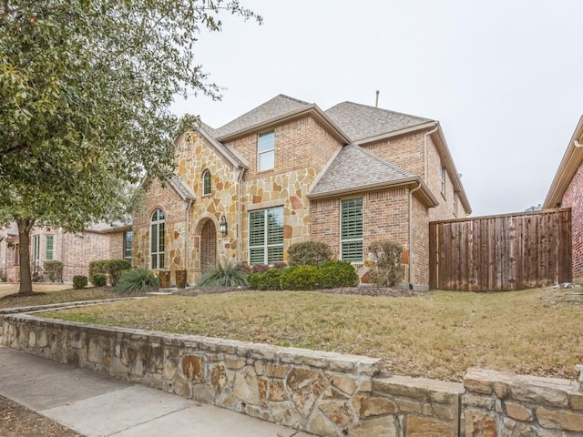 tudor-style house with brick siding, roof with shingles, a front yard, fence, and stone siding