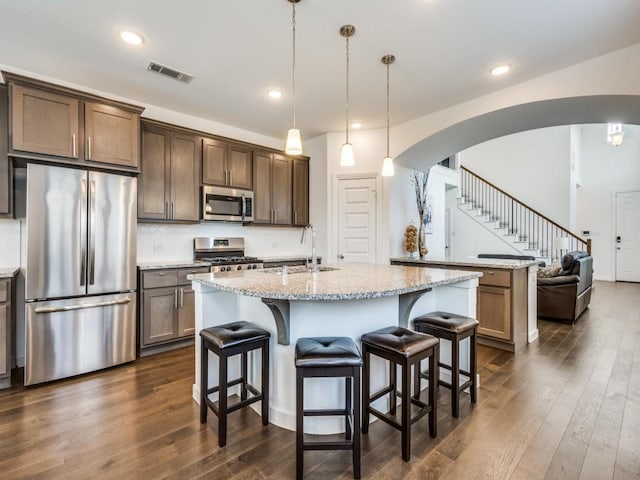 kitchen with light stone counters, a center island with sink, dark hardwood / wood-style flooring, pendant lighting, and stainless steel appliances