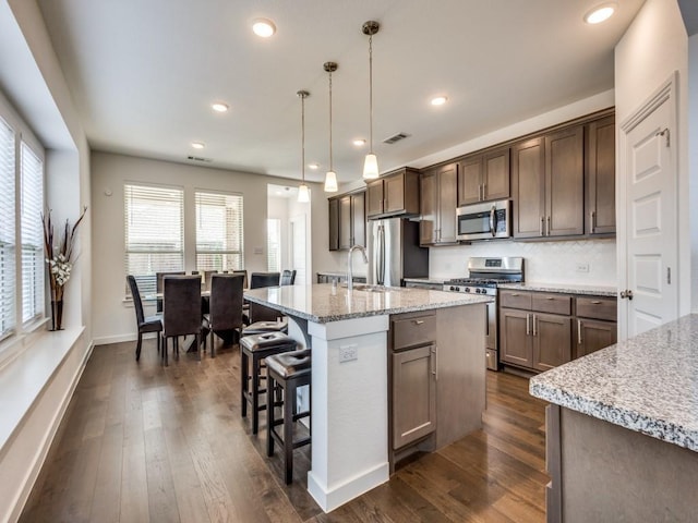 kitchen with decorative light fixtures, a center island with sink, appliances with stainless steel finishes, dark hardwood / wood-style floors, and light stone countertops