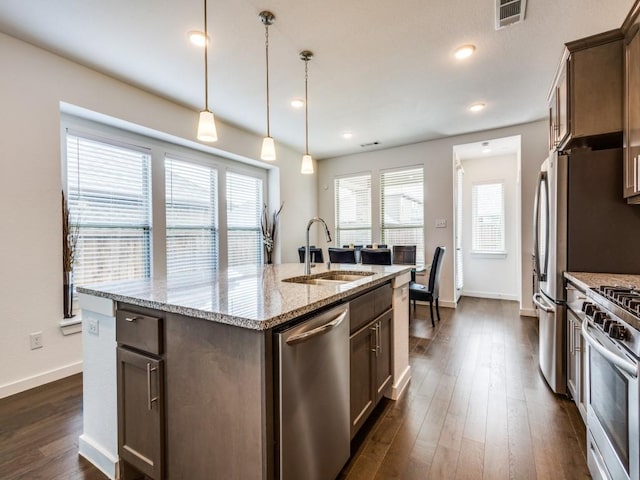 kitchen featuring an island with sink, sink, dark brown cabinetry, stainless steel appliances, and light stone countertops