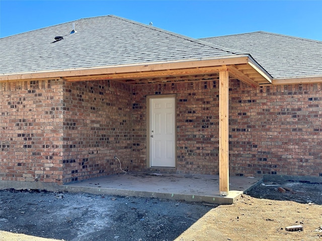 entrance to property with brick siding, roof with shingles, and a patio area