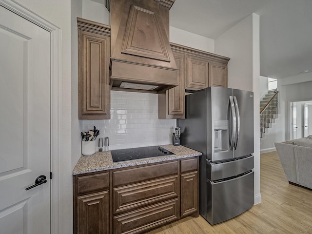 kitchen featuring premium range hood, stainless steel refrigerator with ice dispenser, black electric cooktop, light hardwood / wood-style floors, and backsplash