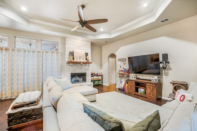 living room featuring a stone fireplace, crown molding, dark hardwood / wood-style floors, a raised ceiling, and ceiling fan