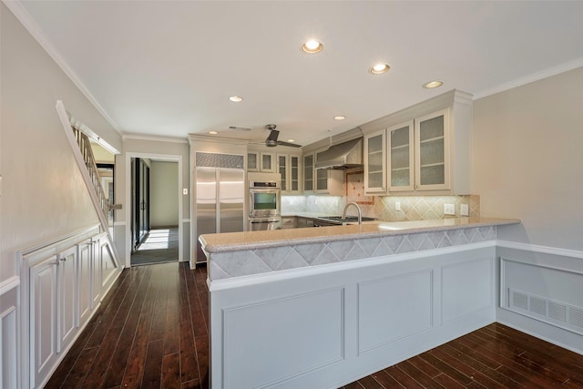 kitchen with built in fridge, decorative backsplash, ornamental molding, kitchen peninsula, and wall chimney range hood