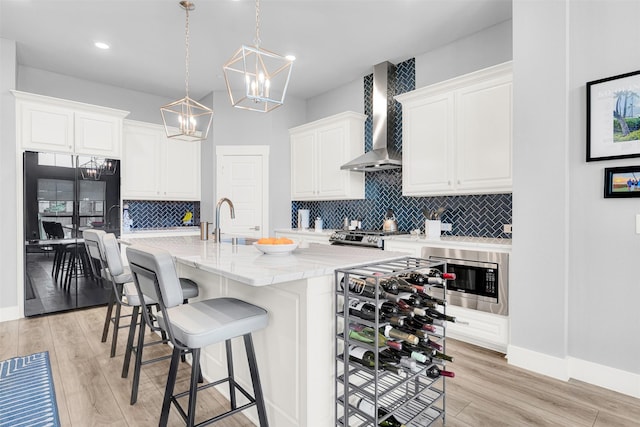 kitchen featuring wall chimney exhaust hood, hanging light fixtures, a center island with sink, appliances with stainless steel finishes, and white cabinets