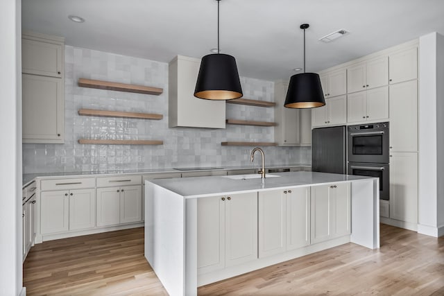 kitchen with sink, a center island with sink, double wall oven, pendant lighting, and white cabinets