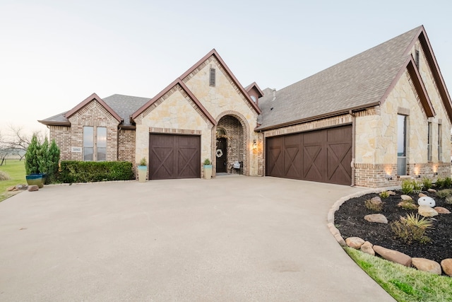 french country inspired facade with stone siding, concrete driveway, brick siding, and an attached garage