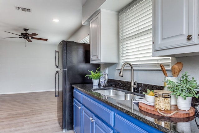 kitchen featuring sink, light hardwood / wood-style flooring, blue cabinetry, black refrigerator, and dark stone countertops