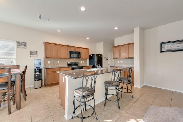 kitchen with a breakfast bar, light tile patterned floors, dark stone counters, a kitchen island with sink, and black appliances