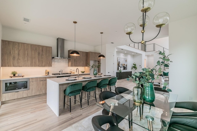 kitchen with black microwave, an island with sink, decorative light fixtures, wall chimney exhaust hood, and light wood-type flooring