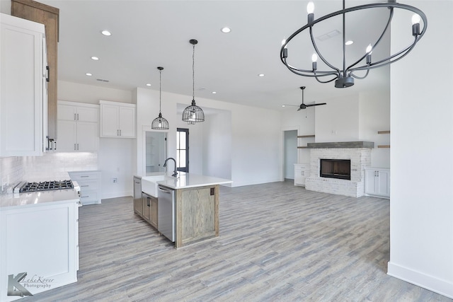 kitchen with a brick fireplace, hanging light fixtures, dishwasher, a kitchen island with sink, and white cabinets