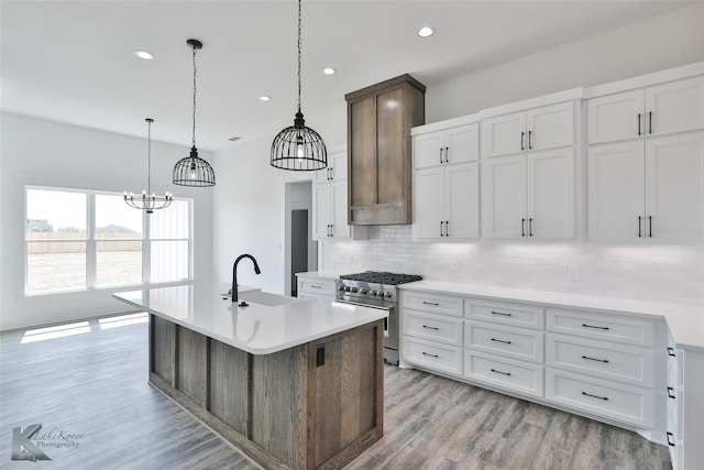 kitchen with decorative light fixtures, an island with sink, sink, white cabinets, and stainless steel range