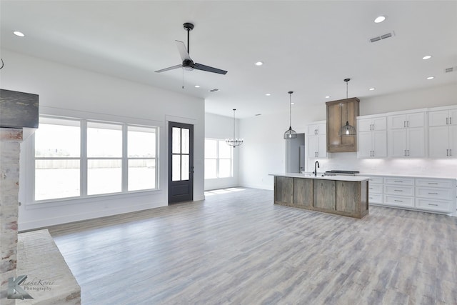 kitchen with pendant lighting, ceiling fan with notable chandelier, white cabinetry, light hardwood / wood-style floors, and a center island with sink