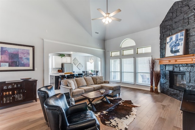 living area with arched walkways, a fireplace, light wood-style flooring, high vaulted ceiling, and ceiling fan with notable chandelier