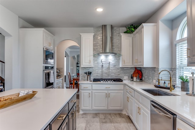 kitchen with appliances with stainless steel finishes, light countertops, wall chimney range hood, white cabinetry, and a sink