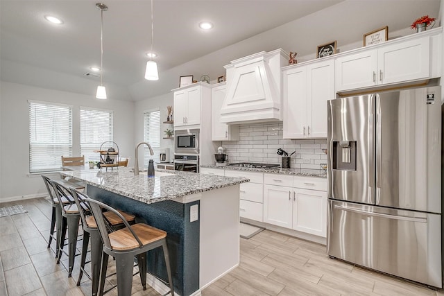 kitchen featuring premium range hood, appliances with stainless steel finishes, an island with sink, white cabinets, and light stone countertops