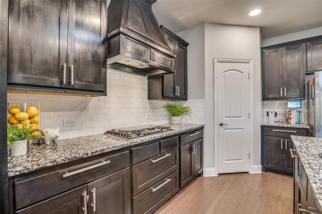 kitchen with dark brown cabinetry, custom exhaust hood, light stone counters, stainless steel appliances, and light hardwood / wood-style floors