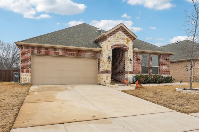 french country inspired facade featuring roof with shingles, brick siding, a garage, stone siding, and driveway