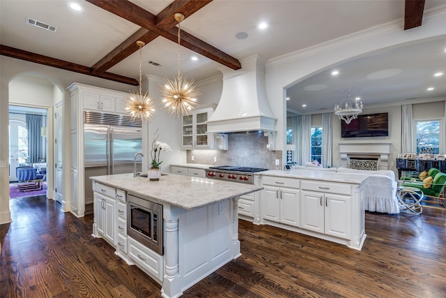 kitchen with premium range hood, built in appliances, dark hardwood / wood-style floors, an island with sink, and white cabinets