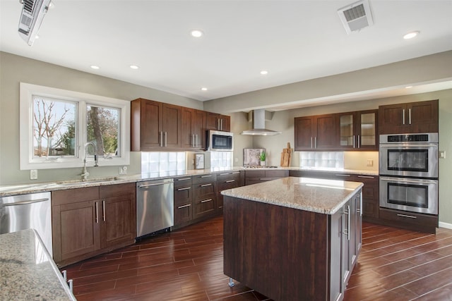 kitchen featuring sink, appliances with stainless steel finishes, a center island, light stone counters, and wall chimney exhaust hood