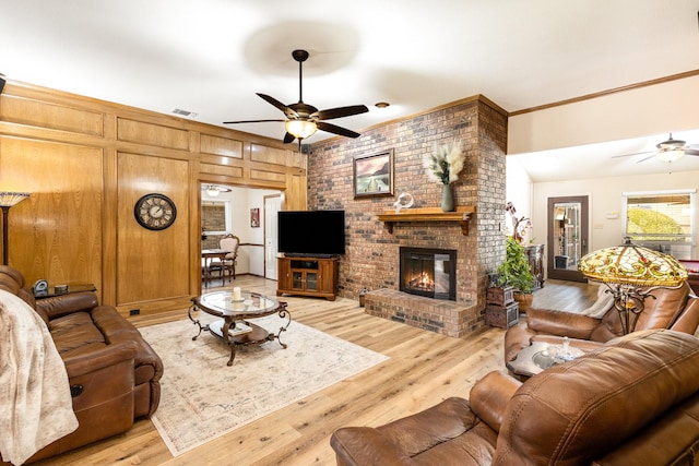 living room featuring crown molding, light hardwood / wood-style flooring, ceiling fan, wooden walls, and a brick fireplace