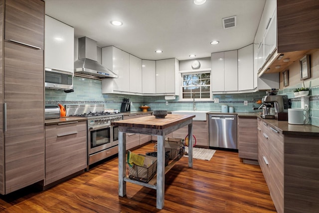 kitchen with sink, appliances with stainless steel finishes, wood-type flooring, white cabinets, and wall chimney exhaust hood