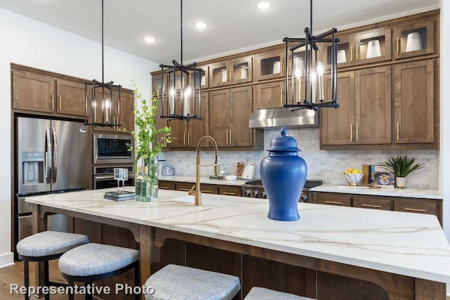 kitchen featuring light stone counters, decorative light fixtures, stainless steel appliances, a kitchen island with sink, and decorative backsplash