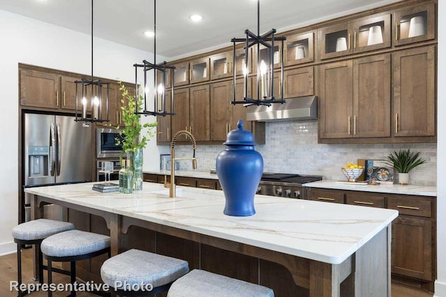 kitchen with hanging light fixtures, an island with sink, stainless steel appliances, light stone countertops, and decorative backsplash