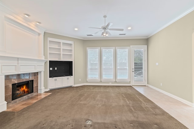 unfurnished living room featuring crown molding, built in features, a tile fireplace, ceiling fan, and light colored carpet