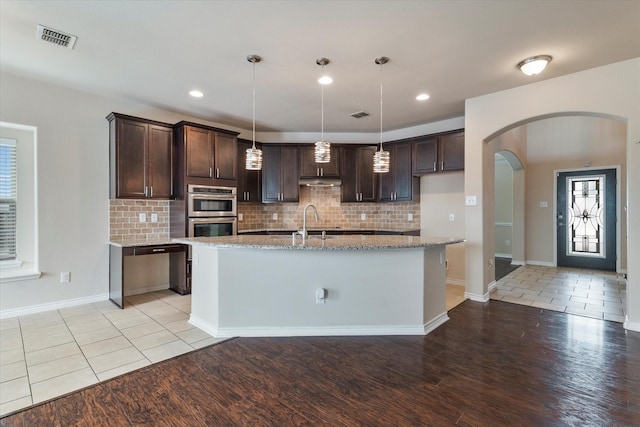 kitchen with dark brown cabinetry, sink, hanging light fixtures, an island with sink, and stainless steel double oven