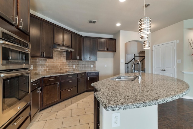 kitchen featuring pendant lighting, sink, stainless steel double oven, dark brown cabinetry, and a center island with sink