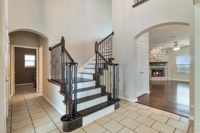 staircase featuring a stone fireplace, tile patterned floors, ceiling fan, and a towering ceiling