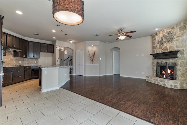 kitchen featuring ceiling fan, a kitchen island with sink, light stone counters, dark brown cabinetry, and a fireplace